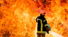 Feuerwehrmann steht vor extrem hohen Flammen und hält Gasflasche in den Händen