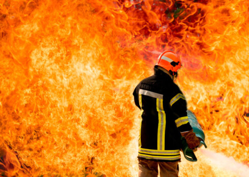 Feuerwehrmann steht vor extrem hohen Flammen und hält Gasflasche in den Händen