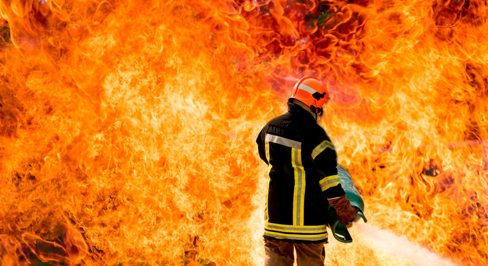 Feuerwehrmann steht vor extrem hohen Flammen und hält Gasflasche in den Händen