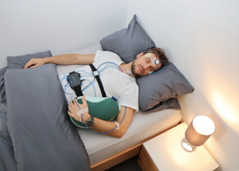 Schlafanalyse in alltagsnaher Situation: Eine Person liegt schlafend auf der Seite in einem Bett und ist an mehrere medizinische Sensoren angeschlossen. Elektroden befinden sich am Kopf, Kabel verlaufen zu einem Messgerät auf der Brust. Ein Gurt um den Oberkörper und ein Sensor am Finger erfassen weitere Daten.