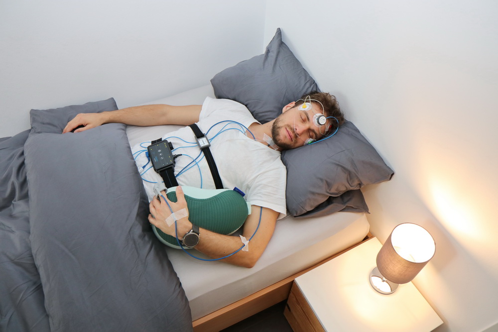 Schlafanalyse in alltagsnaher Situation: Eine Person liegt schlafend auf der Seite in einem Bett und ist an mehrere medizinische Sensoren angeschlossen. Elektroden befinden sich am Kopf, Kabel verlaufen zu einem Messgerät auf der Brust. Ein Gurt um den Oberkörper und ein Sensor am Finger erfassen weitere Daten.