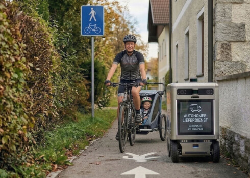 Ein Radfahrer mit Helm fährt auf einem schmalen Weg neben einem Haus und zieht einen Kinderanhänger, in dem ein Kind sitzt. Daneben fährt ein kleiner autonomer Lieferroboter mit der Aufschrift „Autonomer Lieferdienst“. Ein Verkehrszeichen weist den Weg als gemeinsamen Fuß- und Radweg aus.